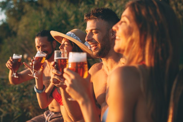 Happy group of young people drinking beer on a dock by the river during the summer sunny day